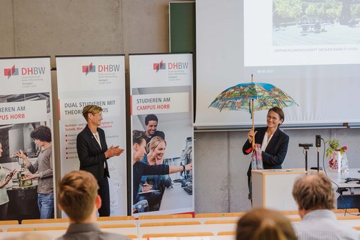 Prof.in Antje Katona überreicht Prof.in Dr. Martina Klärle einen Regenschirm
