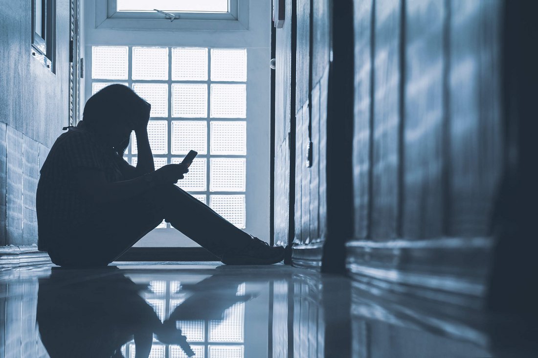 Depressive Frau sitzt allein in einem Hausflur und schaut auf ihr Smartphone.