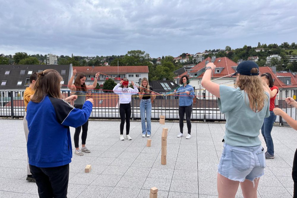 Die Teilnehmenden stehen im Kreis und halten jeweils eine Schnur in den Händen, die in der Mitte des Kreises zusammenlaufen. 