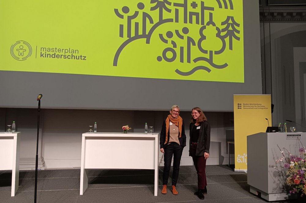 Prof. Dr. Anja Teubert und Milena Buhl von der DHBW Stuttgart stehen auf einer Bühne, im Hintergrund ist auf einer Leinwand ein großes Plakat "masterplan kinderschutz" zu sehen.