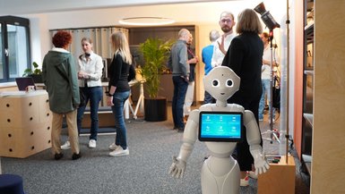 Eröffnung des Studios41 Roboter Pepper steht im Vordergrund, um die Gäste zu begrüßen, im Hintergrund informieren sich Besucher*innen über das Studio41.