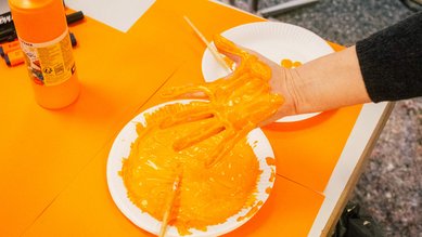 Großaufnahme des Tellers mit orangefarbener Fingerfarbe, in die eine Hand getunkt und in die Kamera gehalten wird.