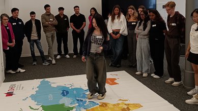 Internationale Studierende steht auf einer auf dem Boden ausgebreiteten Weltkarte, die weiteren internationalen Studierenden gruppieren sich um diese Karte herum.
