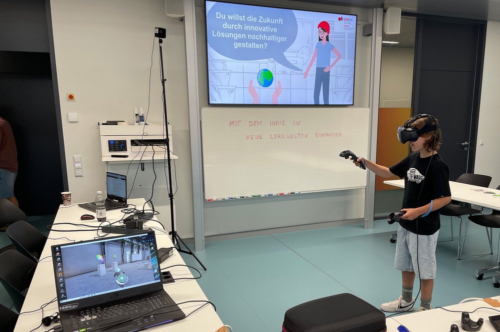 Jugendlicher probiert eine VR-Brille aus. Im Hintergrund wird ein Bild an die Wand projiziert, auf dem steht: "Du willst die Zukunft durch innovative Lösungen nachhaltiger gestalten?"