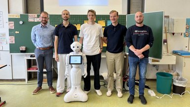 Von den Studierenden extra für dieses Setting programmiert: Pepper-Roboter des ZDT hilft im Unterricht.(von li. nach re.: Erich Heumüller, Niklas Ruch, Maximilian Ganzert, Fynn Deitermann, Bastian Weippert) Gruppenbild mit dem Roboter Pepper im Klassenzimmer vor der Tafel.