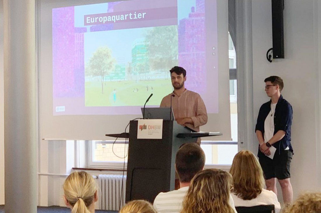 Einer der beiden Studierenden steht am Mikrofon, im Hintergrund sieht man den Ausschnitt einer Präsentation.