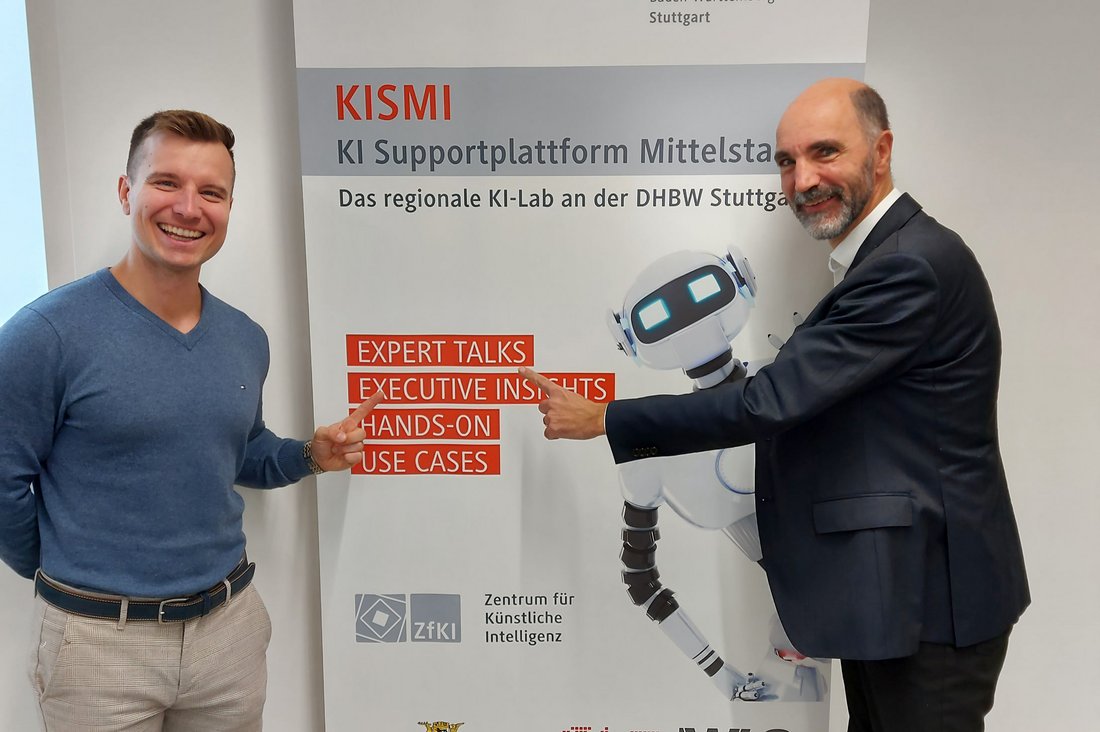 Dr. Christian Tutschku vom Fraunhofer IAO und Prof. Dr. Dirk Reichhardt von der DHBW Stuttgart vor dem Plakat der Veranstaltung