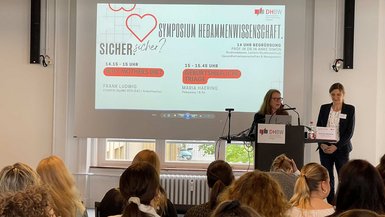 Symposium Hebammenwissenschaft der DHBW Stuttgart am 11. Mai 2023 Vortragende am Rednerpult, im Hintergrund ist das Programm des Symposiums zu sehen.