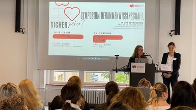 Vortragende am Rednerpult, im Hintergrund ist das Programm des Symposiums zu sehen.