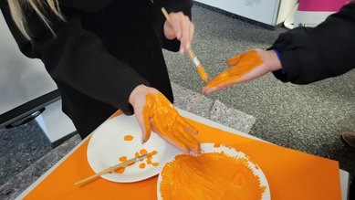 Besucher*innen des Infostands der Fakultät Sozialwesen am 25.11.2025 bemalen ihre Hände mit orangefarbener Fingerfarbe, um einen Handabdruck zu hinterlassen.