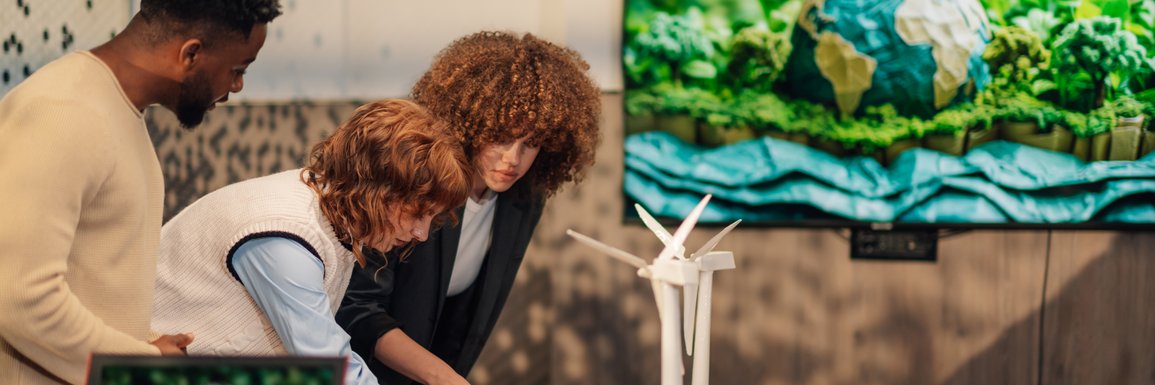 Personen arbeiten an einem Schreibtisch mit Miniaturen von Windrädern und TV mit Nachhaltigkeitsmotiv