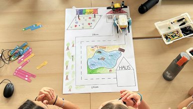 Ein bunt angemalter Spielplan liegt auf einem Tisch. Auf dem Spielplan steht ein kleiner Roboter, um den Tisch herum sitzen Schüler*innen einer Grundschule.