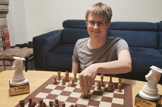 Tobias Kölle sitzt vor einem Schachbrett, neben dem links und rechts jeweils einen Pokal stehen.