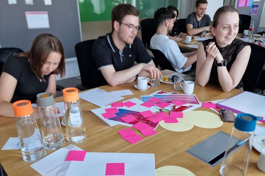 Teilnehmende sitzen an einem Tisch und arbeiten. Auf dem Tisch liegen u. a. Blöcke und Klebezettel.