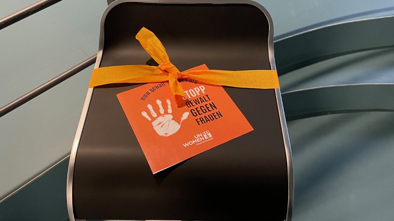 Stuhl mit orangefarbenem Kreppband und einem DIN-A-4-Plakat Stopp Gewalt gegen Frauen. 