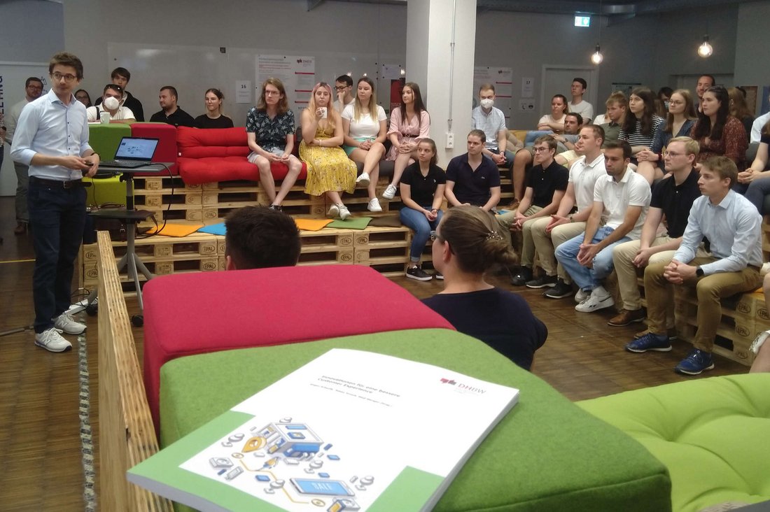 Gruppenfoto mit Studierenden, die auf Palettenmöbeln im Halbkreis um einen Vortragenden sitzen.