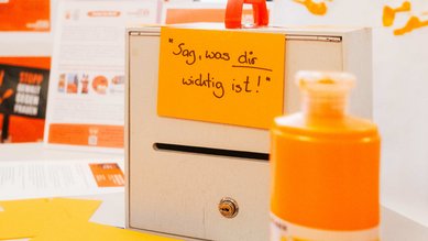 Großaufnahme des "Kummerkastens", in den Besucher*innen des Standes ihre auf orangefarbenen Karteikarten geschriebenen Anmerkungen einwerfen konnten.