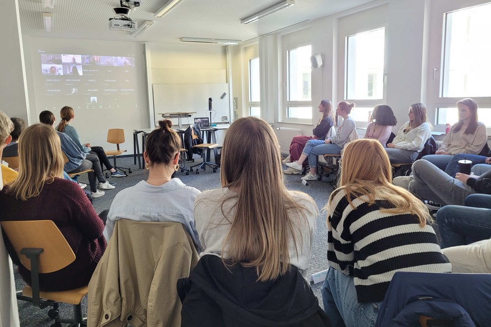 Studierende sitzen im Halbkreis und schauen nach vorne auf eine Leinwand, auf der die Lesung übertragen wurde.