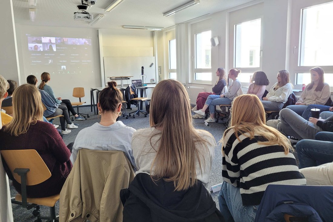 Studierende sitzen im Halbkreis und schauen nach vorne auf eine Leinwand, auf der die Lesung übertragen wurde.