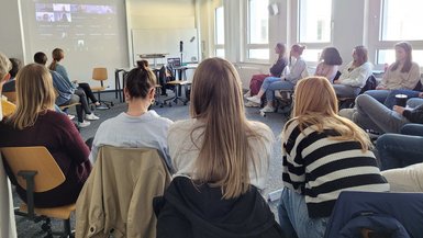 Studierende sitzen im Halbkreis und schauen nach vorne auf eine Leinwand, auf der die Lesung übertragen wurde.