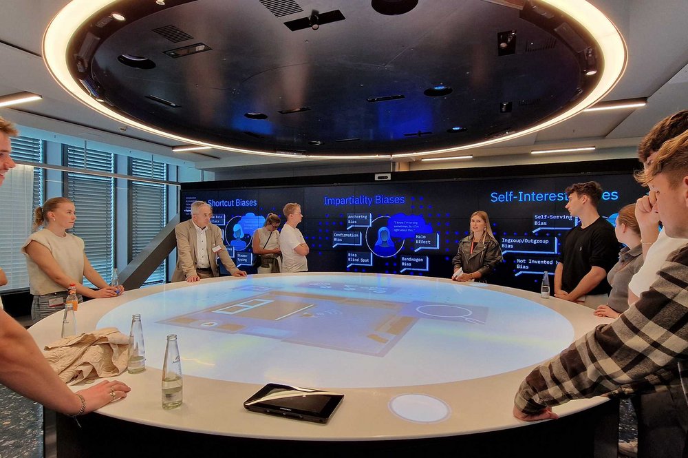 Teilnehmende stehen an einem runden Tisch mit einer großen Projektionsfläche in der Mitte.