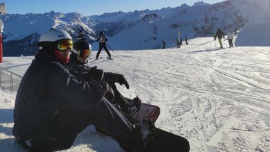 Zwei Teilnehmende der Exkursion sitzen im Schnee und schauen in Richtung der Ski-Piste.
