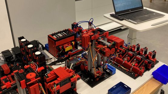 Im Vordergrund steht eine Modellfabrik von Fischertechnik mit Hochregallager zur Veranschaulichung der vollautomatischen Abwicklung von Aufträgen.